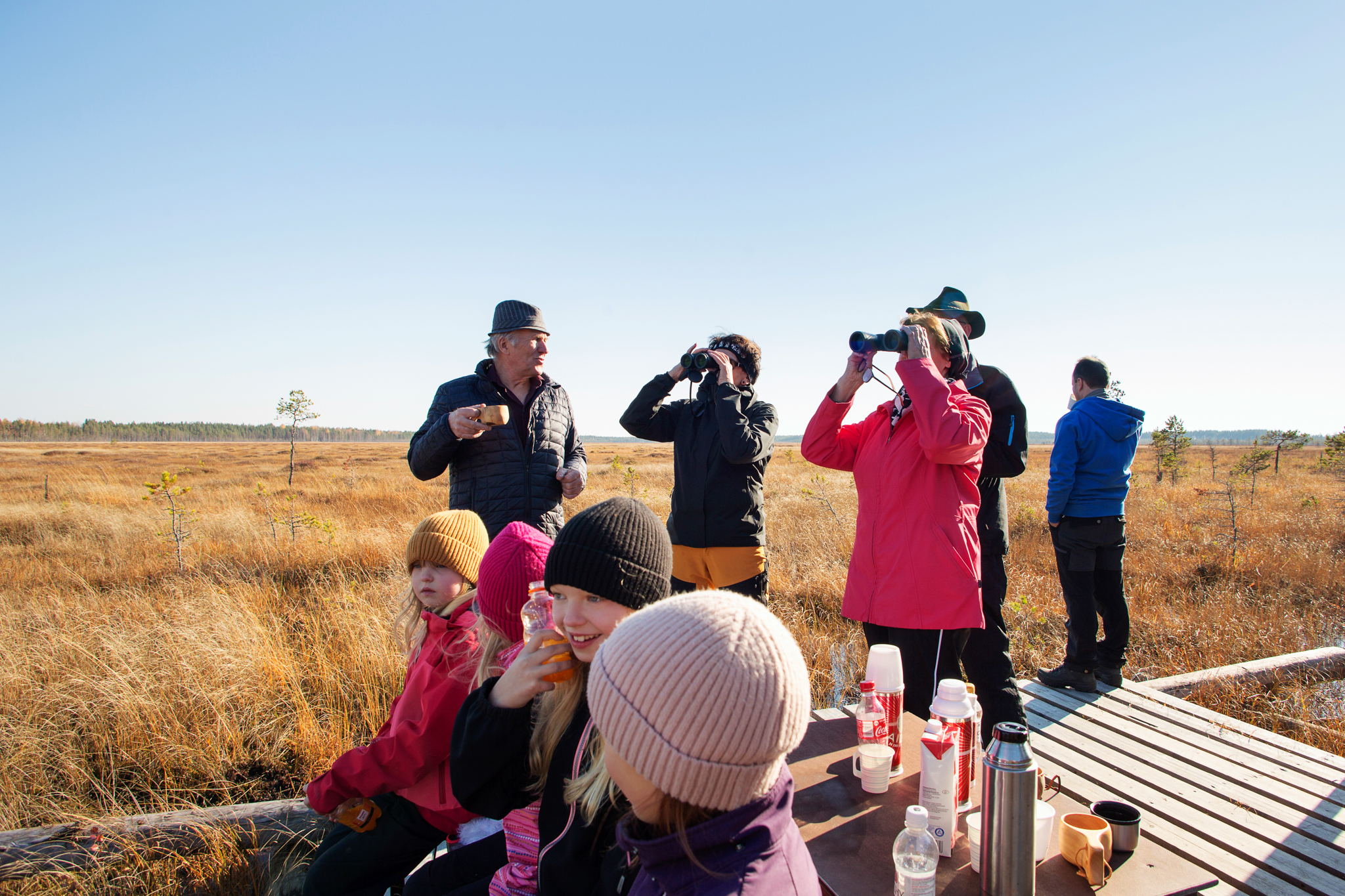 Joukko retkeilijöitä pitkospuureitin päässä olevalla tarkkailutasanteella syömässä eväitä, osa katselee kiikareilla suomaisemaa.