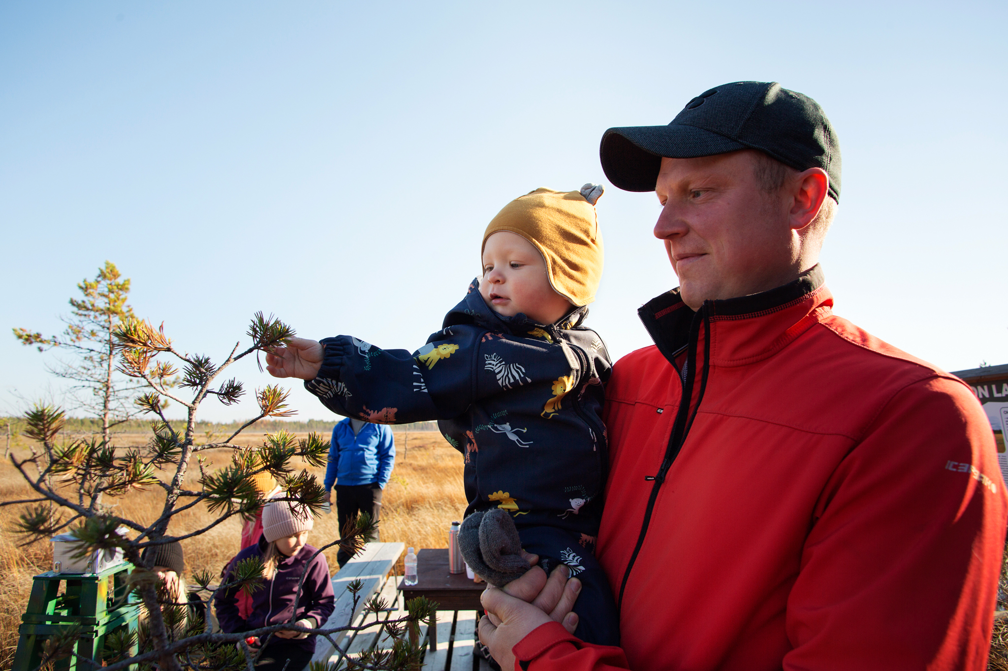 Taustalla suomaisema, etualalla Pertti Tanskanen pieni lapsi sylissään, taustalla joukko retkeilijöitä eväitä syömässä.