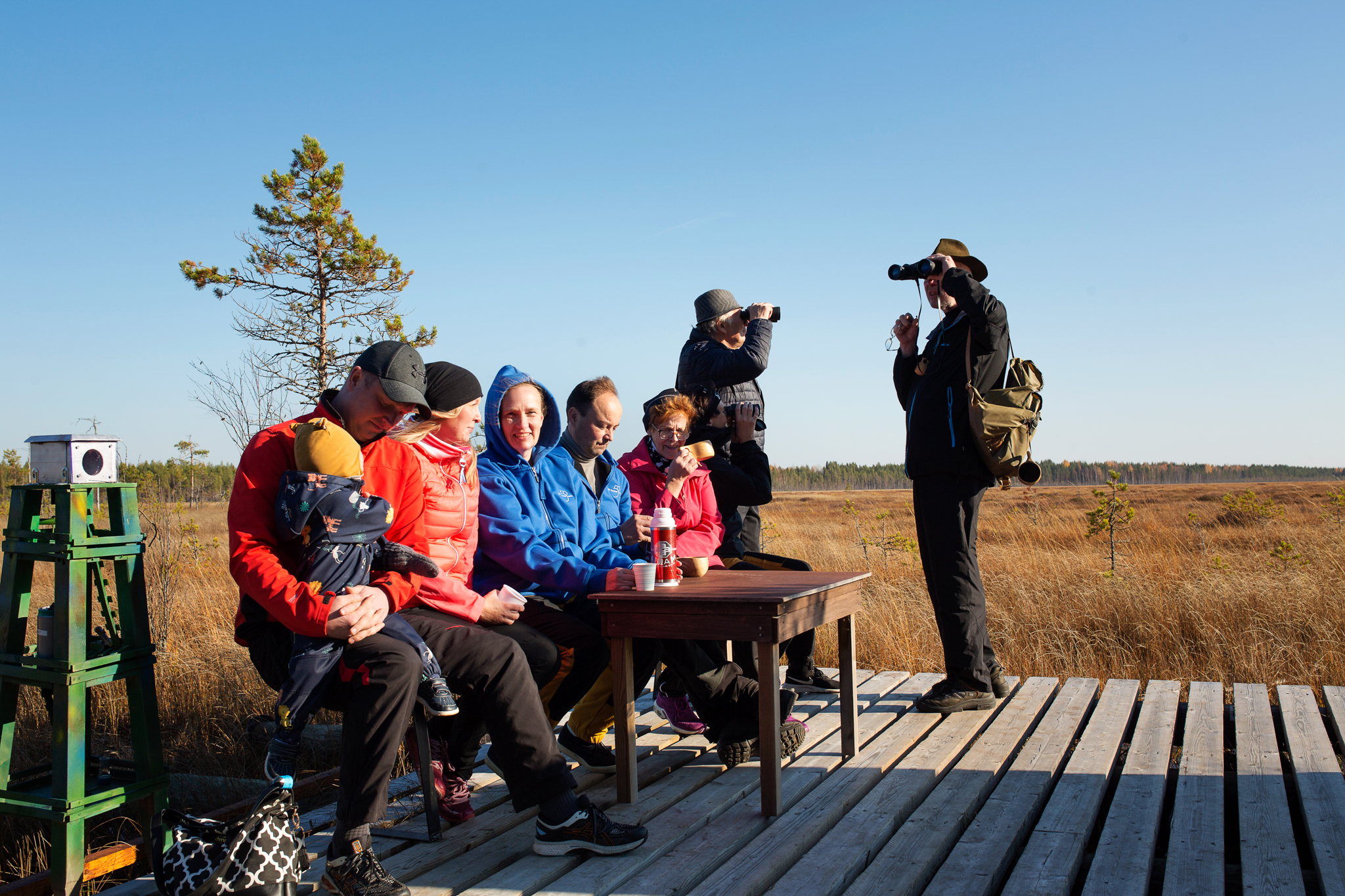 Joukko retkeilijöitä pitkospuureitin päässä olevalla tarkkailutasanteella syömässä eväitä, osa katselee kiikareilla suomaisemaa.