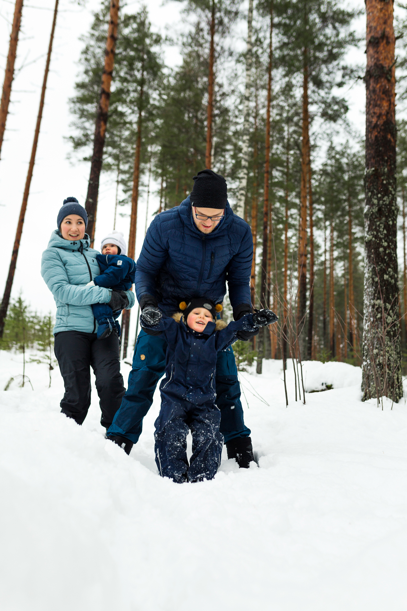 Nainen ja mies kahden lapsen kanssa lumisessa mäntymetsässä.