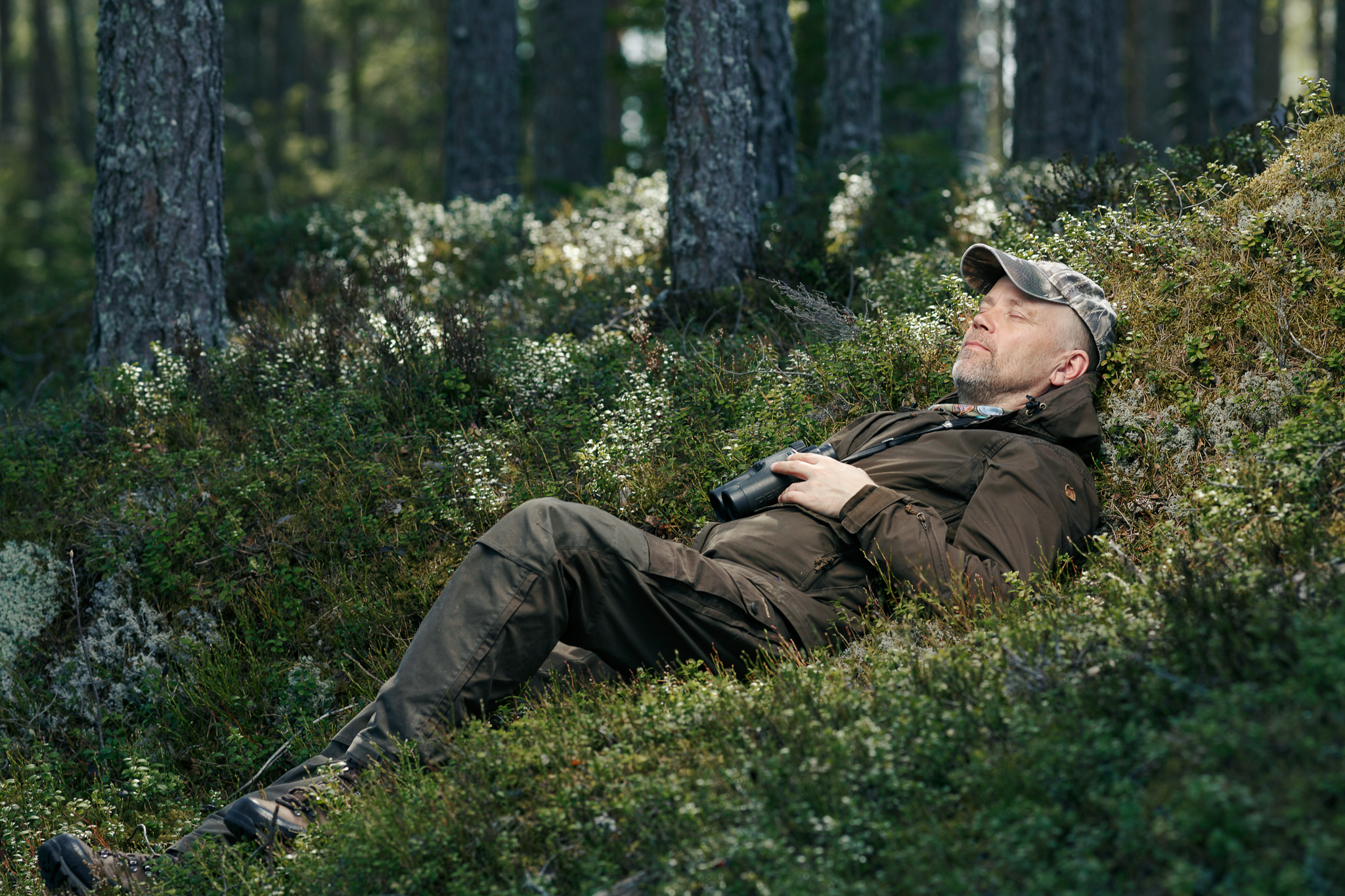 Kuvassa mies makaamassa silmät kiinni sammaleisessa maastossa, kädessänä hänellä on kiikarit. Taustalla mäntymetsää.