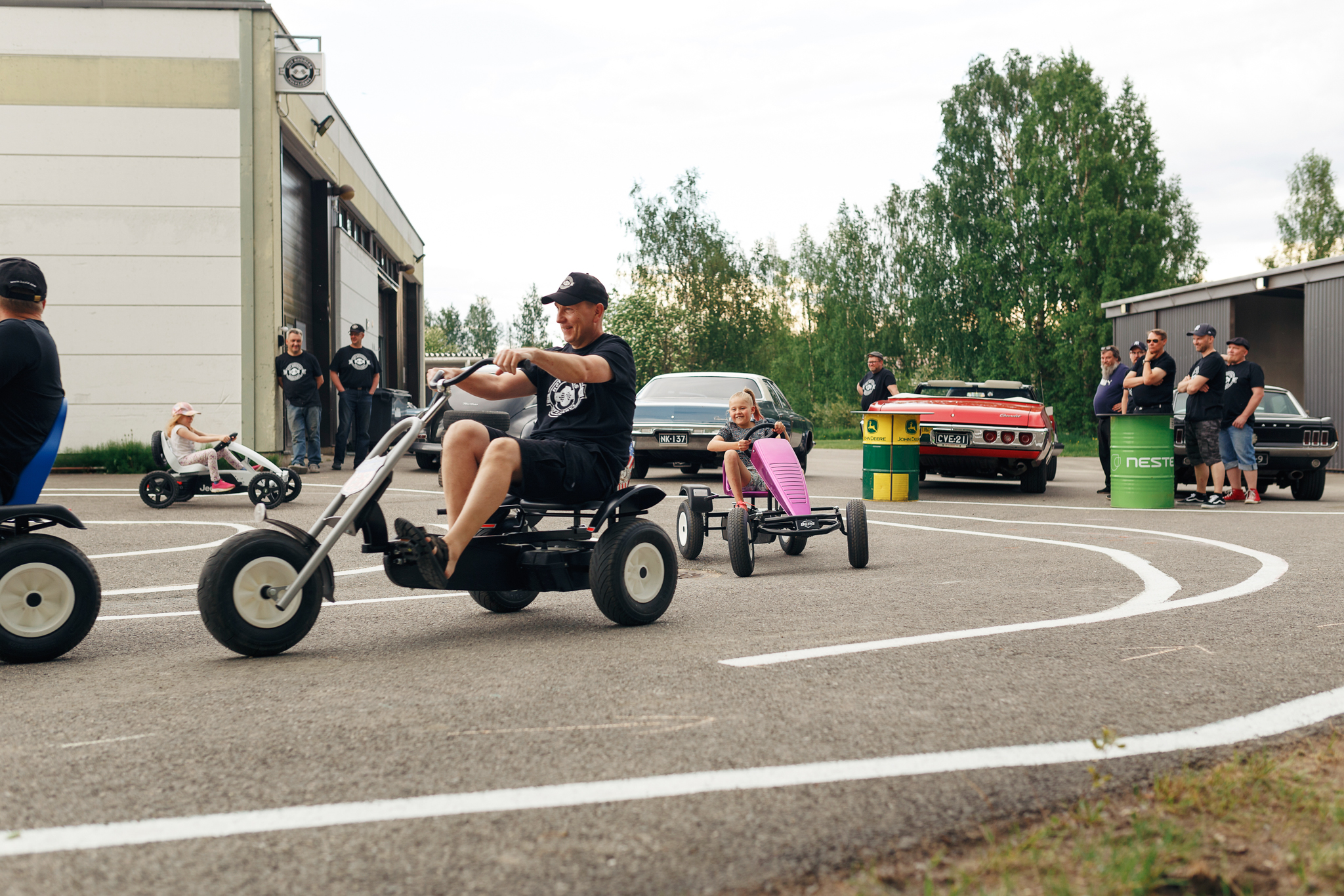 Liikennepuisto autohallin vieressä. Mika Rieppo ja Olli-Peka Karttunen ajavat kilpaa polkuautoilla kahden pienen tytön kanssa, muut yhdistyksen jäsenet seuraavat sivusta.